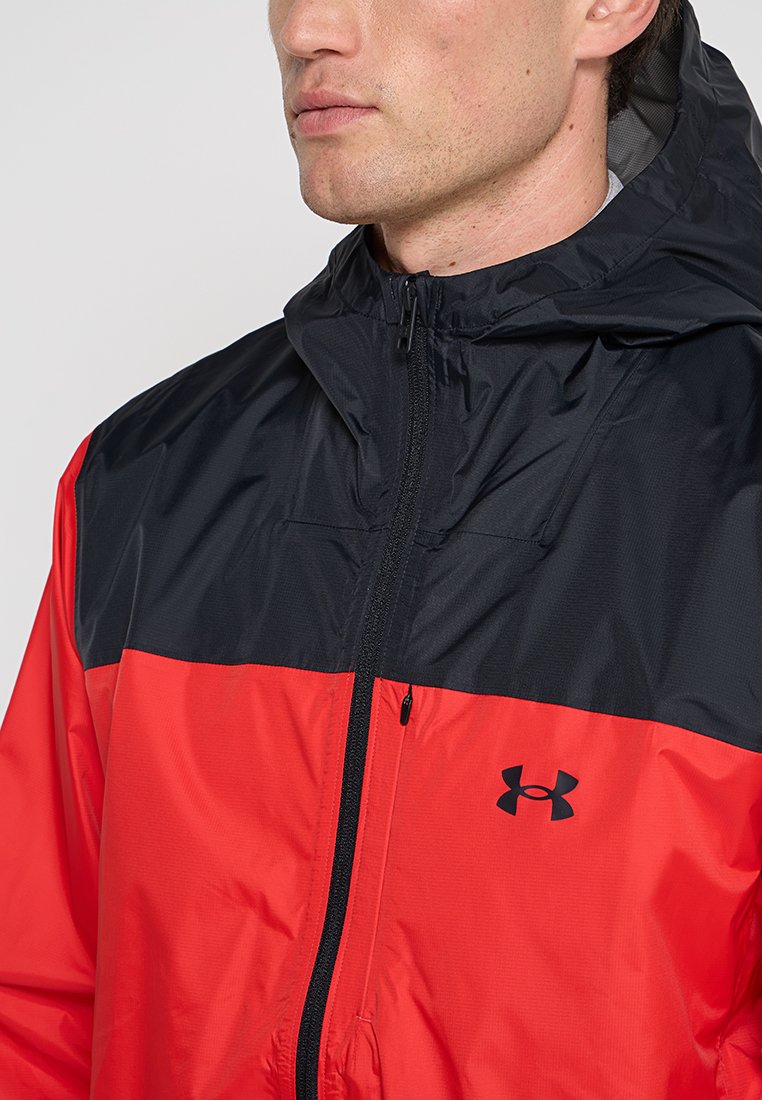 Uomo che indossa una giacca a zip con cappuccio rossa e nera con il logo Under Armour sul petto, rivolto a sinistra.