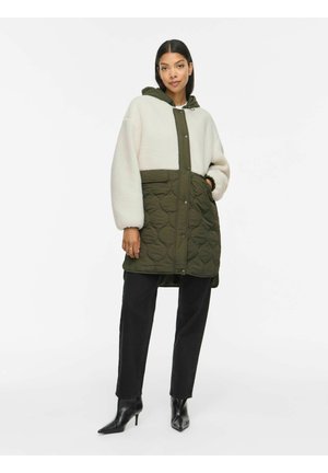 Vrouw draagt een tweekleurige jas met een witte fleece bovenkant en doorgestikte olijfgroene onderkant, zwarte broek en zwarte laarzen met hak tegen een witte achtergrond.