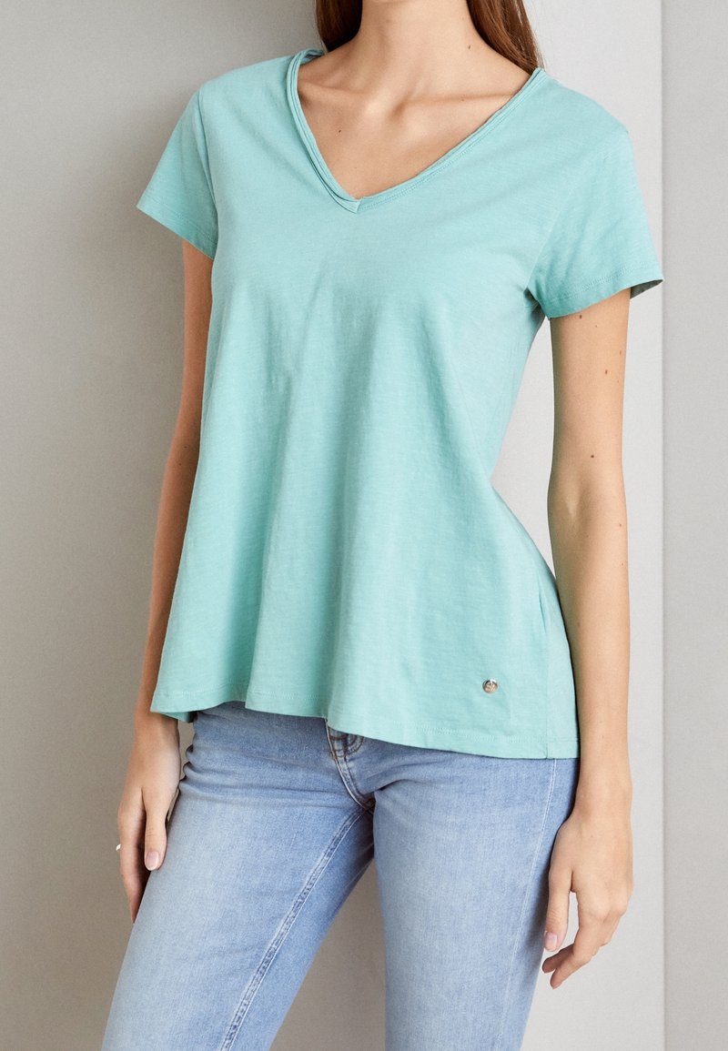 T-shirt à col en V de couleur turquoise clair avec manches courtes, fabriqué en tissu doux. Présente une coupe décontractée avec un subtil accent métallique à l'ourlet.