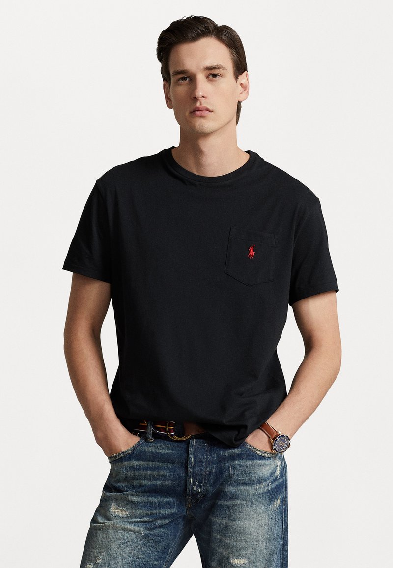 Black cotton t-shirt featuring a round neckline and a chest pocket, accented with a small red embroidered logo. Worn with distressed blue jeans.