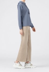 Blå huvtröja med luva och avslappnad passform, kombinerad med beige vida byxor och vita kraftiga sneakers. Mjukt tyg och enkel design.