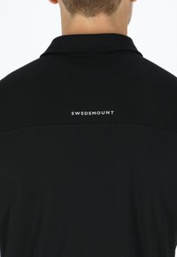 Svart pikétröja med texturerad tyg, kort krage och en vit "SWEDEMOUNT"-logotyp på den övre delen av ryggen. Enkelt design, elegant silhuett.