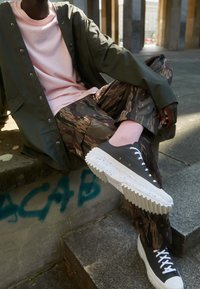 Svarta sneakers med räfflade vita sulor, kamouflage-mönstrade byxor och en rosa tröja under en olivgrön jacka, sittande på stentrappor.