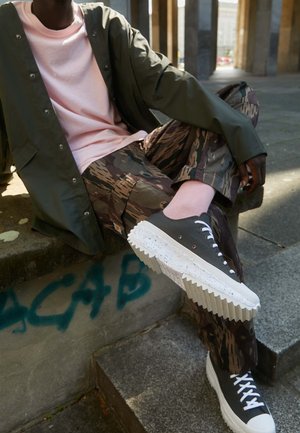 Schwarze Sneakers mit strukturierten weißen Sohlen, camouflage-musterte Hose und ein pinkes Shirt unter einer olivgrünen Jacke, sitzend auf Steinstufen.
