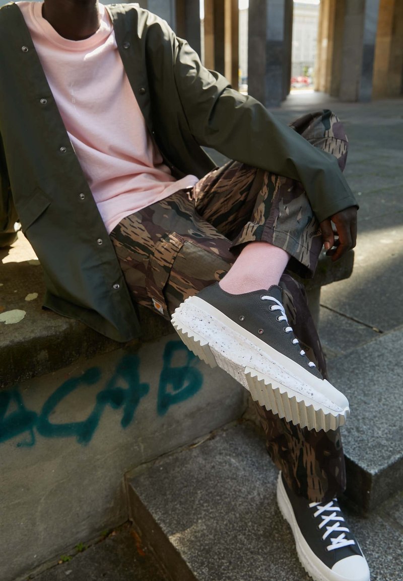 Svarta sneakers med räfflade vita sulor, kamouflage-mönstrade byxor och en rosa tröja under en olivgrön jacka, sittande på stentrappor.