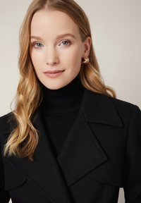 Czarny wełniany płaszcz z wyrazistym kołnierzem, noszony na dopasowanym czarnym golfie. Włosy są długie i faliste, ozdobione złotymi kolczykami w kształcie kół.