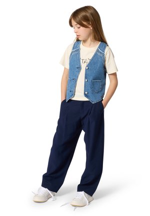 Denim spijkervest met een lichte wassing, knoopsluiting en twee voorzakken, gecombineerd met een crèmekleurig T-shirt en marineblauwe wijde broek. Witte sneakers.