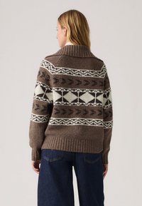 Rjav pleten pulover z visokim ovratnikom, ki ima bele in temne vzorce, rebrast rob in manšete, oblečen z temno denim hlačami.