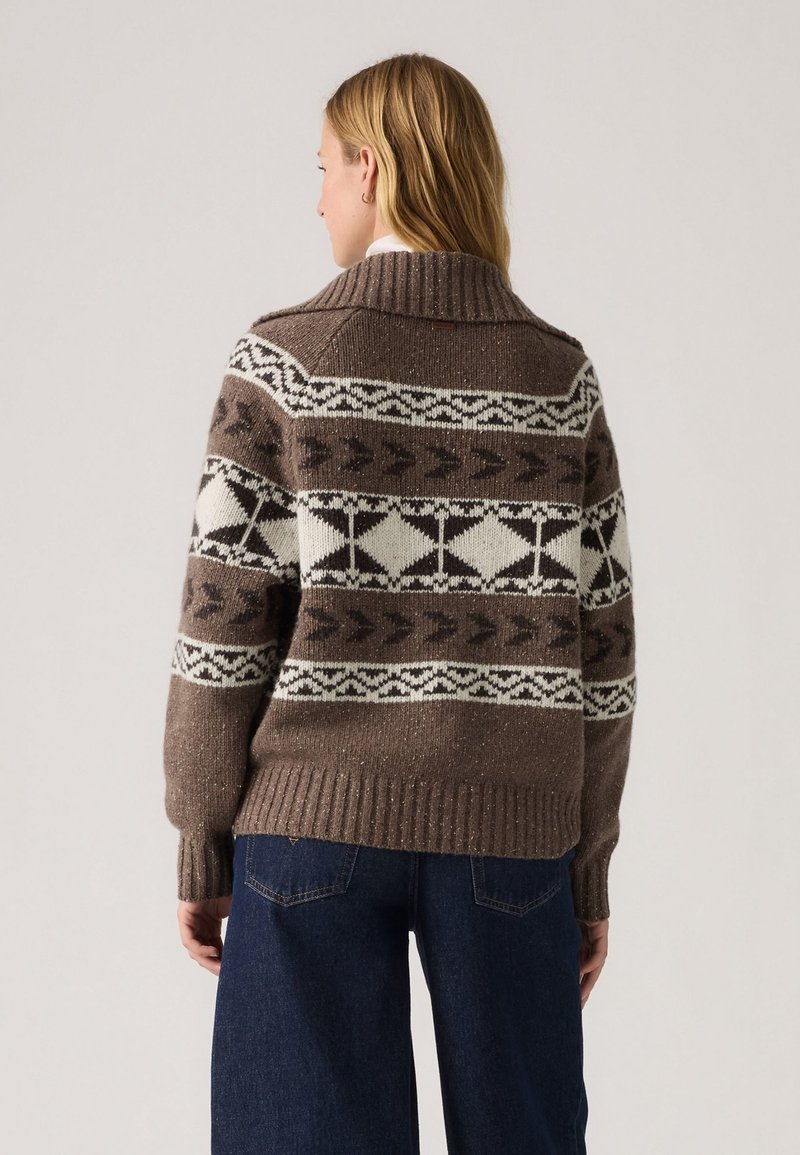 Brun strikket sweater med høj krave, prydet med hvide og mørke mønstre, ribbet kant og manchetter, båret med mørke jeans.