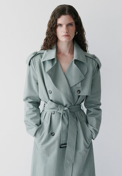 Kvinde med krøllet brunt hår iført lys grøn trenchcoat med bælte, stående med hænderne i lommerne mod en ensfarvet baggrund.