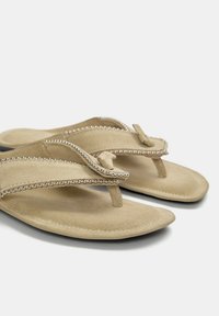 Beiga sandaler med en mjuk mockakvalitet, med en tåpostdesign, dekorerade med silvriga nitar längs remmarna. Platt sula.