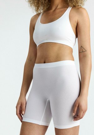 Kobieta ubrana w biały, prążkowany sportowy biustonosz i dopasowane, wysokie szorty kolarskie, pokazująca prosty tatuaż w kształcie trójkąta na lewej ręce.