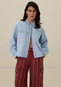 Hellblaues Jeansjackett mit zwei Brusttaschen, Knöpfen vorne und lockerer Passform; kombiniert mit rostfarben gestreiften, weiten Hosen.