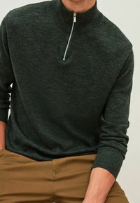 Tmavě zelený svetr na zip s vysokým límcem, vyrobený z texturovaného pleteného materiálu, s jemným logem na levé straně hrudi.