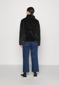 Giacca in pelliccia sintetica nera con colletto alto, abbinata a pantaloni in denim blu a gamba larga e sneakers nere alte. Texture liscia, vestibilità casual.