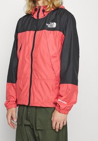 Červeno-černá větrovka s předním zipem, elastickými manžetami a nenápadným logem. Lehká, hladká textura s kapucí.