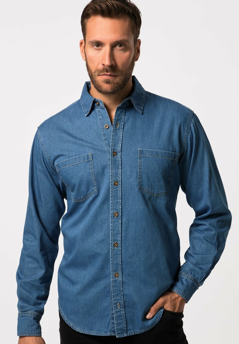 Camisa de mezclilla en azul medio con mangas largas, dos bolsillos en el pecho, cierre frontal con botones y botones marrones en contraste. Textura suave, corte clásico.