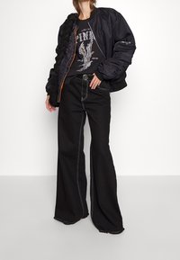 Černá bomber bunda s prošívanými detaily na ramenou, spárovaná s černým tričkem s grafikou orla a černými vysokými rozšířenými džíny s bílým švem.