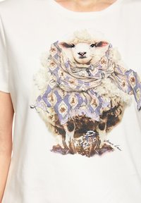 White t-shirt featuring a graphic of a sheep wearing a patterned purple scarf with embellishments. The design includes a signature at the bottom.