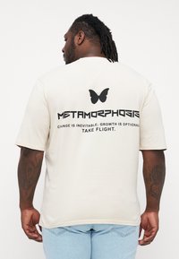 Cream-colored t-shirt made of soft fabric, featuring a large black butterfly graphic and text on the back in bold typography.