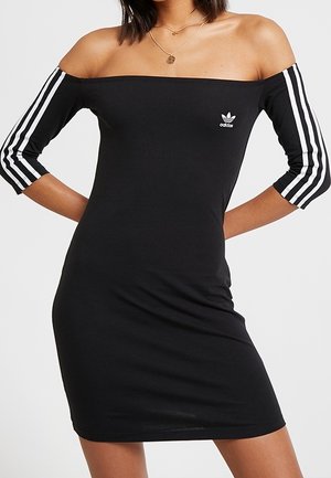 Kobieta ubrana w czarną sukienkę Adidas z odsłoniętymi ramionami, z trzema białymi paskami na rękawach oraz małym logo na piersi.