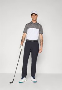 Golfoutfit met een grijs-wit poloshirt, marineblauwe broek, witte schoenen met blauwe accenten en een golfclub in de hand.