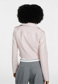 Gekürzte rosa Kunstlederjacke mit Schulterklappen und vertikalen Nähten, kombiniert mit grauen Hosen und einem weißen Hemd darunter.