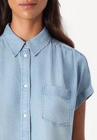 Kvinne som har på seg en lyseblå denim kortermet skjorte med knapper, som har en brystlomme, krage og oppbrettede ermekanter.