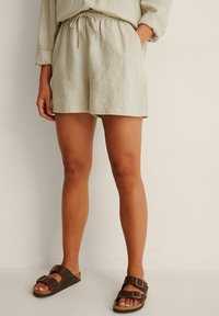 Personne portant un short en lin beige avec des poches avant et des sandales marron à double lanière, debout contre un mur clair uni.