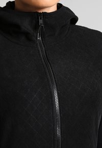 Zwarte zip-up hoodie van textuurstof met gewatteerd materiaal. Heeft een hoge kraag en een opvallende rits aan de voorkant.