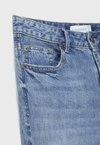 Close-up of light blue denim jeans front pocket with copper rivets and a visible brand label inside the waistband.