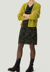 Zielona kardiganowa sweter z przędzy na czarnej sukience w żółte liściaste wzory. Czarne kozaki do kolan. Przezroczyste czarne rajstopy.