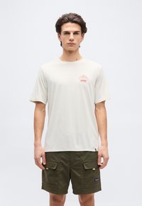 Νέος άνδρας στέκεται με το πρόσωπο προς τα εμπρός φορώντας μια μπεζ κοντομάνικη μπλούζα με μικρό κόκκινο σχέδιο και χακί σορτς cargo.