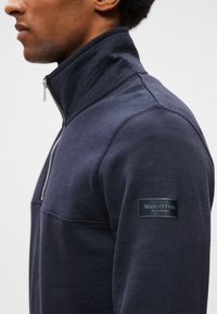 Giacca a zip in pile blu scuro con colletto alto, texture a coste e tasca laterale. Presenta un'applicazione con logo sulla manica superiore.