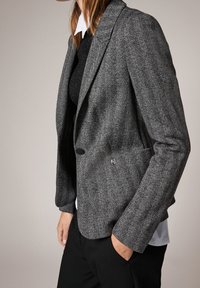 Blazer à motif chevrons noir et blanc en tissu texturé, avec un col structuré, une fermeture à deux boutons et une coupe ajustée.