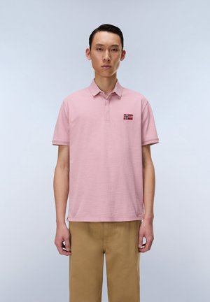 Giovane uomo che indossa una polo rosa con un piccolo logo a forma di bandiera e pantaloni beige, in piedi davanti a uno sfondo chiaro e semplice.