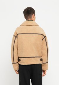 Beige, fuzzy shearling jakke med brune læderdetaljer og syninger. Har en bred krave og en løs pasform. Bagview vist.