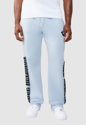 Mężczyzna w jasno niebieskich spodniach dresowych z czarnym pionowym napisem po bokach, białych tenisówkach i białej koszulce, stojący na jednolitym tle.