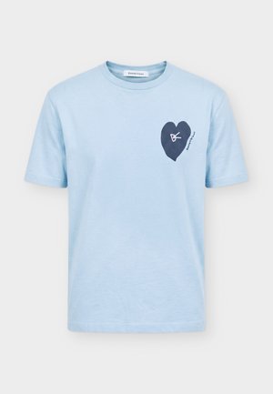 Giovane uomo con capelli corti e ricci che indossa una maglietta azzurro chiaro con una grafica di cuore scuro sul petto e pantaloni neri.