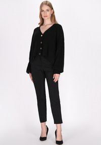 Zwarte oversized gebreide cardigan met een V-hals, voorzien van grote gouden knopen, gecombineerd met slim fit zwarte broeken en zwarte hoge hakken.
