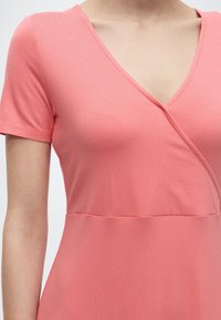 Frau trägt ein korallenrosa kurzärmeliges Kleid mit V-Ausschnitt und Wickeldetail an der Brust, abgebildet von der Schulter bis zur Taille.