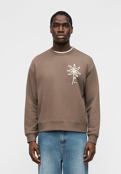 Homme portant un sweat-shirt marron avec un motif de fleur blanche sur la poitrine et un jean bleu, debout devant un fond clair uni.