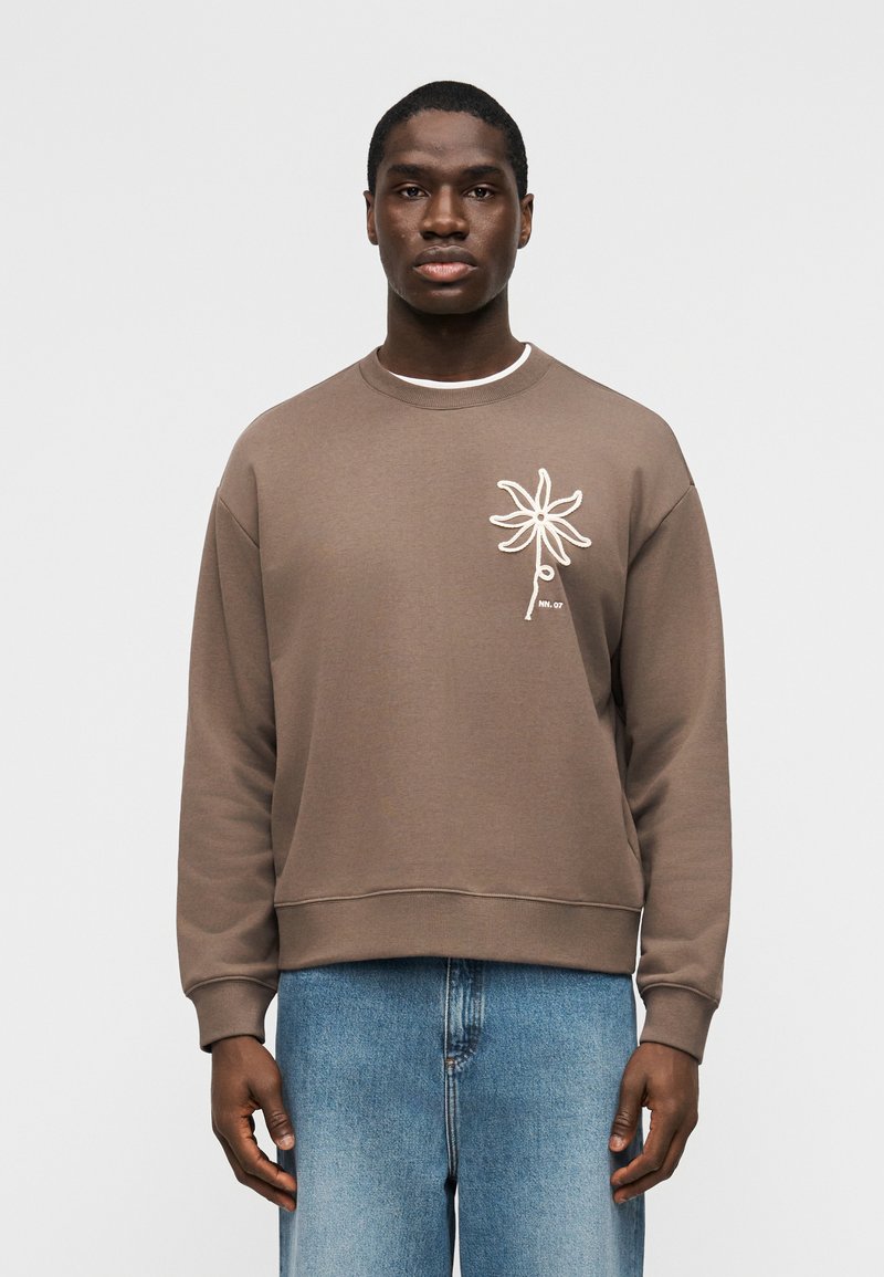 Άνδρας που φοράει καφέ φούτερ με λευκό σχέδιο λουλουδιού στο στήθος και μπλε τζιν, στέκεται μπροστά σε απλό φωτεινό φόντο.