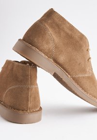 Bottines en daim marron avec semelles en caoutchouc beige, coutures visibles et fermeture à lacets sur fond blanc.