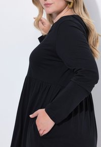 Robe noire à manches longues en tissu doux, présentant une coupe décontractée, une taille froncée et des poches latérales. Texture lisse avec une silhouette fluide.