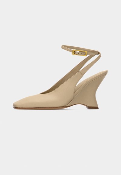 Μπεζ γόβα με μυτερό άκρο και πέδιλο τύπου slingback με κεκλιμένο τακούνι τύπου σφήνα και λουράκι αστραγάλου με χρυσή αγκράφα σε λευκό φόντο.