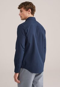 Man draagt een donkerblauw hemd met lange mouwen en lichtgrijze broek, gezien van achteren met het hoofd iets naar links gedraaid.