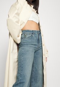 Abrigo largo beige claro sobre un top corto blanco y jeans de mezclilla de lavado medio. Los jeans presentan un parche con logo y un diseño ajustado.
