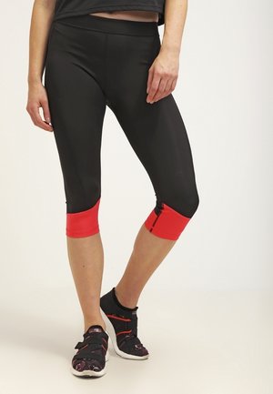 Kvinde iført sorte og røde cropped leggings og sorte atletiske sko med velcro-remme, stående mod en ensfarvet baggrund.