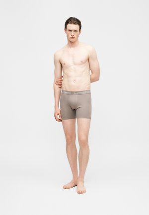 Jaunas vyras stovi basas, dėvi šviesiai rudos spalvos Calvin Klein trumpikes prieš paprastą baltą foną.